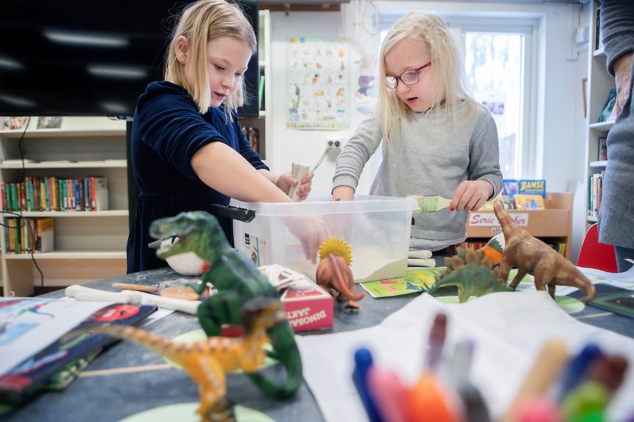 Två barn tittar i en plastlåda som står på ett bord. På bordet står det dinosaurier och färgpennor.